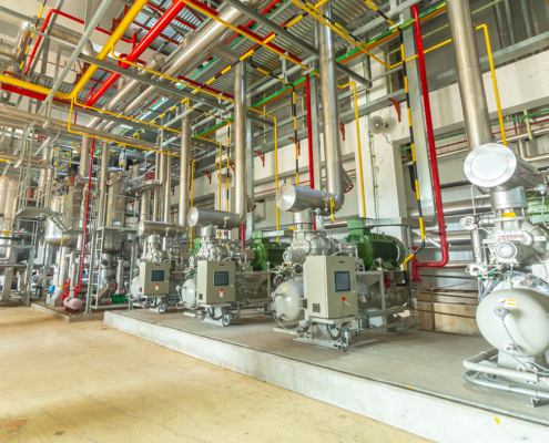 Industrial refrigeration plant room with compressors, stainless-steel pipework, control panels, and color-coded utility lines.